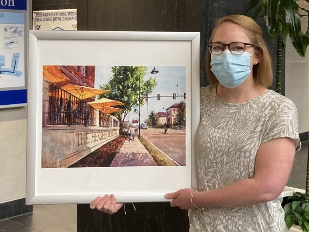 Lauren Talhelm proudly stands holding a framed waercolor painting titled “Afternoon in Hershey” by Brienne Brown 