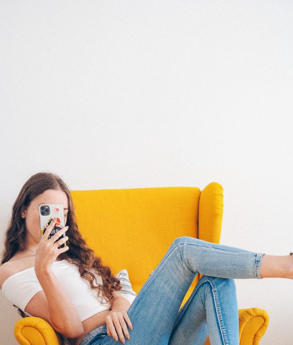 woman in white top and jeans on yellow arm chair holding smartphone