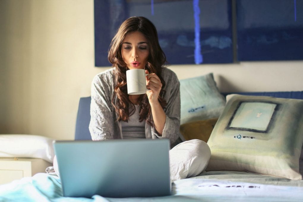 Woman sitting on bed with computer and mug of hot beverage