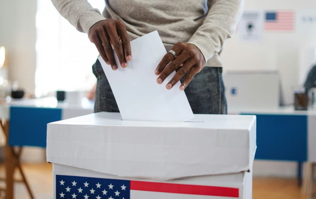 A Black man putting a paper ballot into a ballot box emblazoned with an American flag.