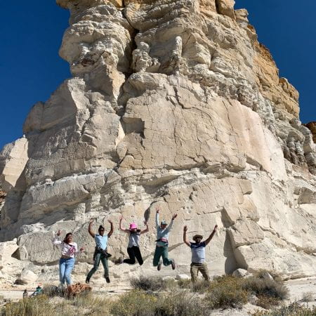 Five geologists leap for joy in front of a white-tan sandstone outcrop under a sunny clear blue sky