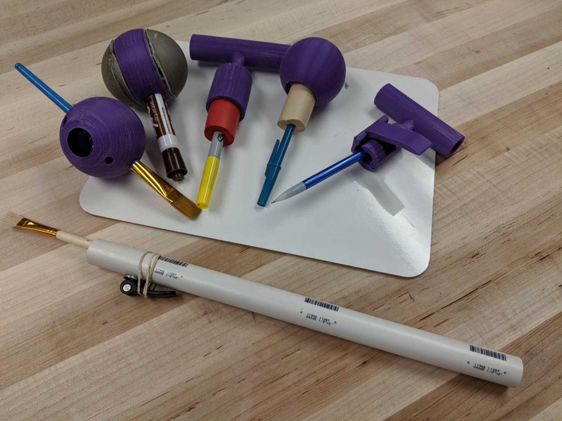 Photo of six art tools (2 paintbrushes, a dry erase marker, a sharpie, a pen, and a pencil) all with prototype handles attached to the ends of them